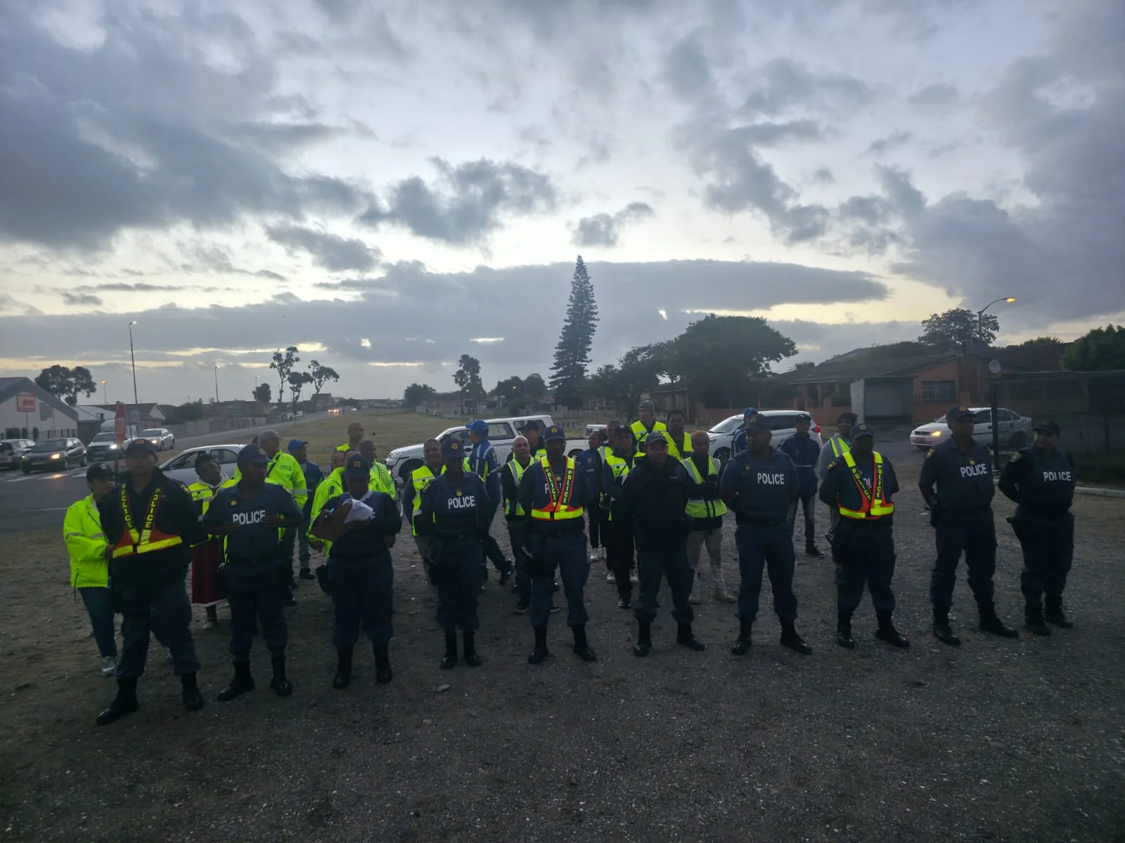 A large outdoor gathering of neighbourhood-watch participants and uniformed safety personnel at dusk.